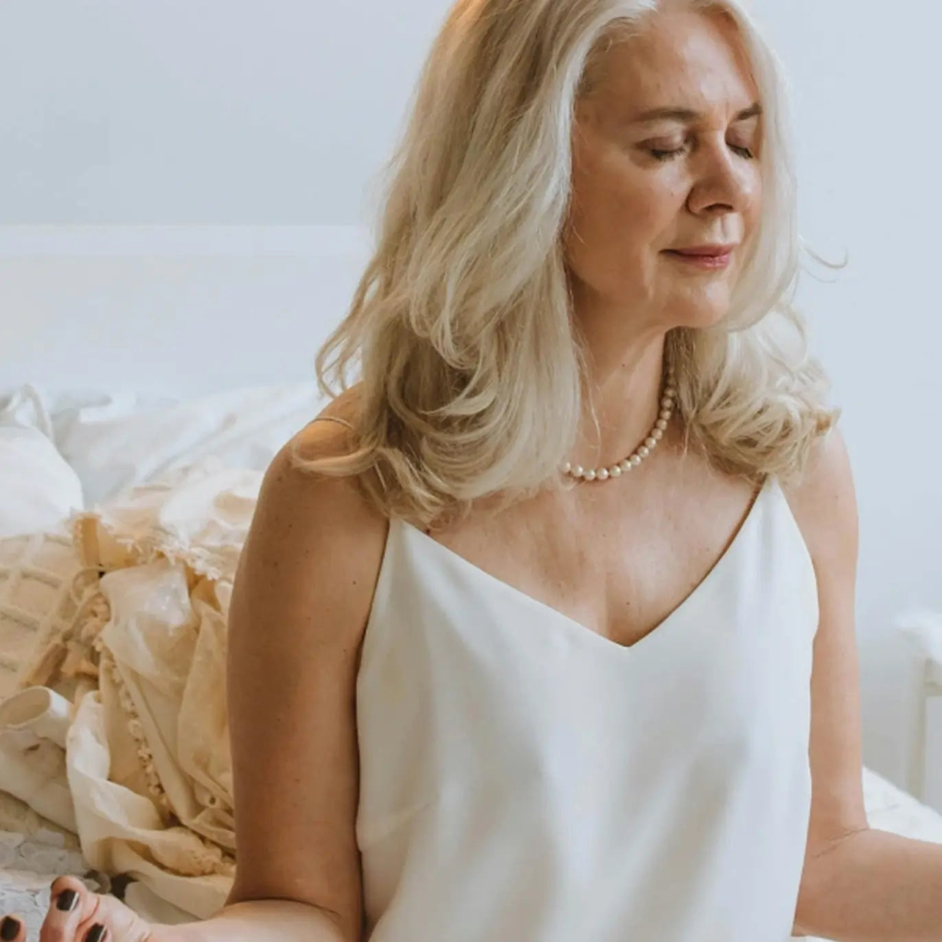 Woman wearing a white sleeveless top with a pearl necklace in a softly lit room.