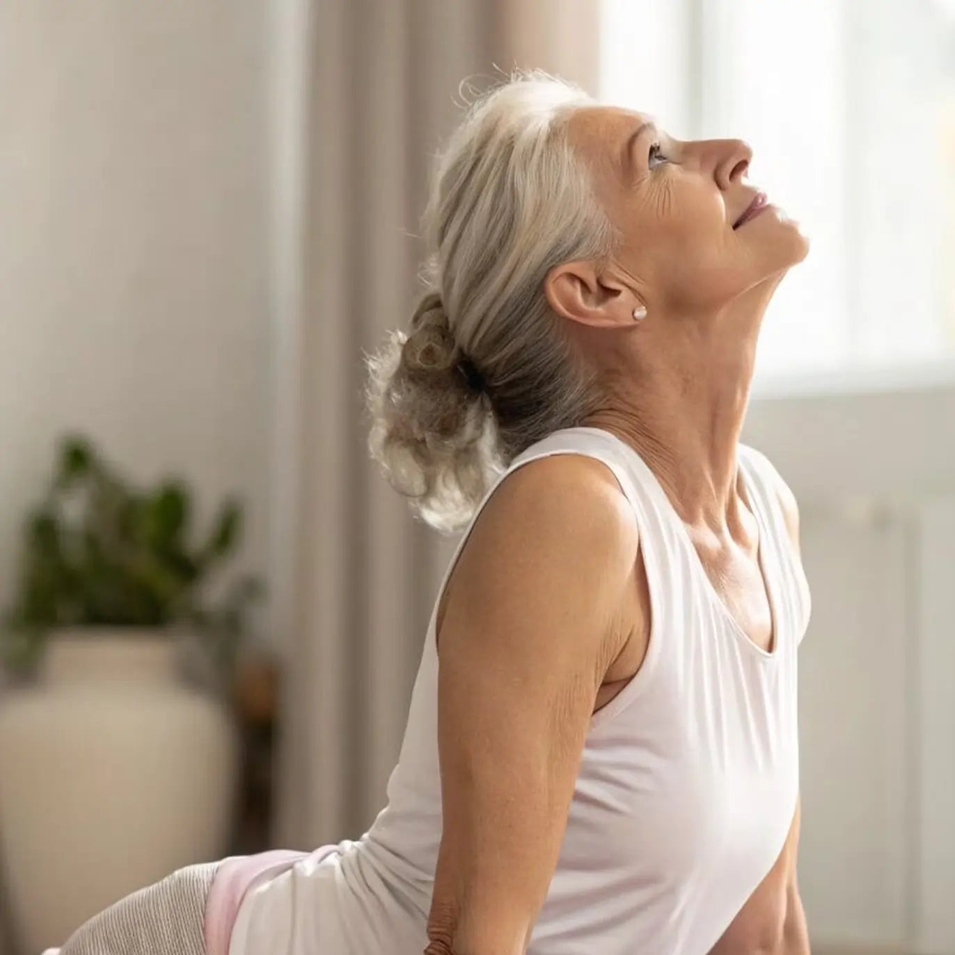 Woman in a white tank top stretching in a bright room with plants.