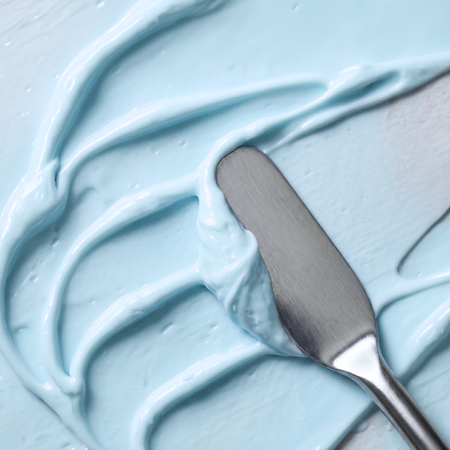 A close-up of thick, light blue cream or gel with a metal spatula resting on top, showing smooth swirled texture.