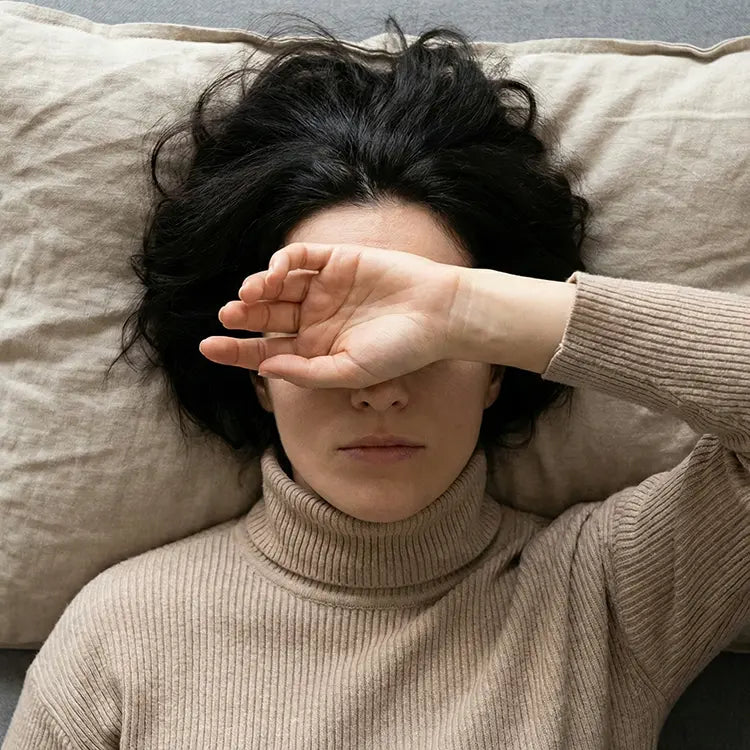 Person wearing a beige sweater lying on a pillow with one hand covering their face.